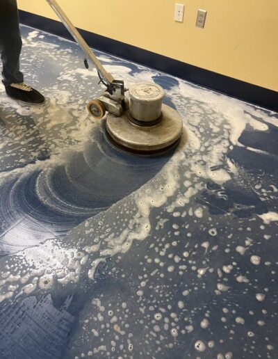 Technician using a floor buffing machine to scrub a blue tile floor with foaming cleaner